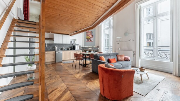 A spacious living area with a herringbone wood floor, a staircase with a glass railing, a grey sofa with orange cushions, and a view of the city through large windows. - Fenêtre sur cour - Studio pour 2 (Lyon)