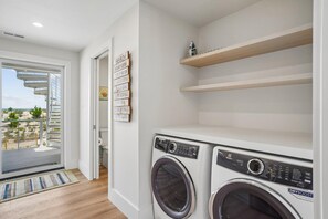 A laundry room with two front-loading washing machines, a wooden shelf, and a door leading to a balcony with a view of the beach.