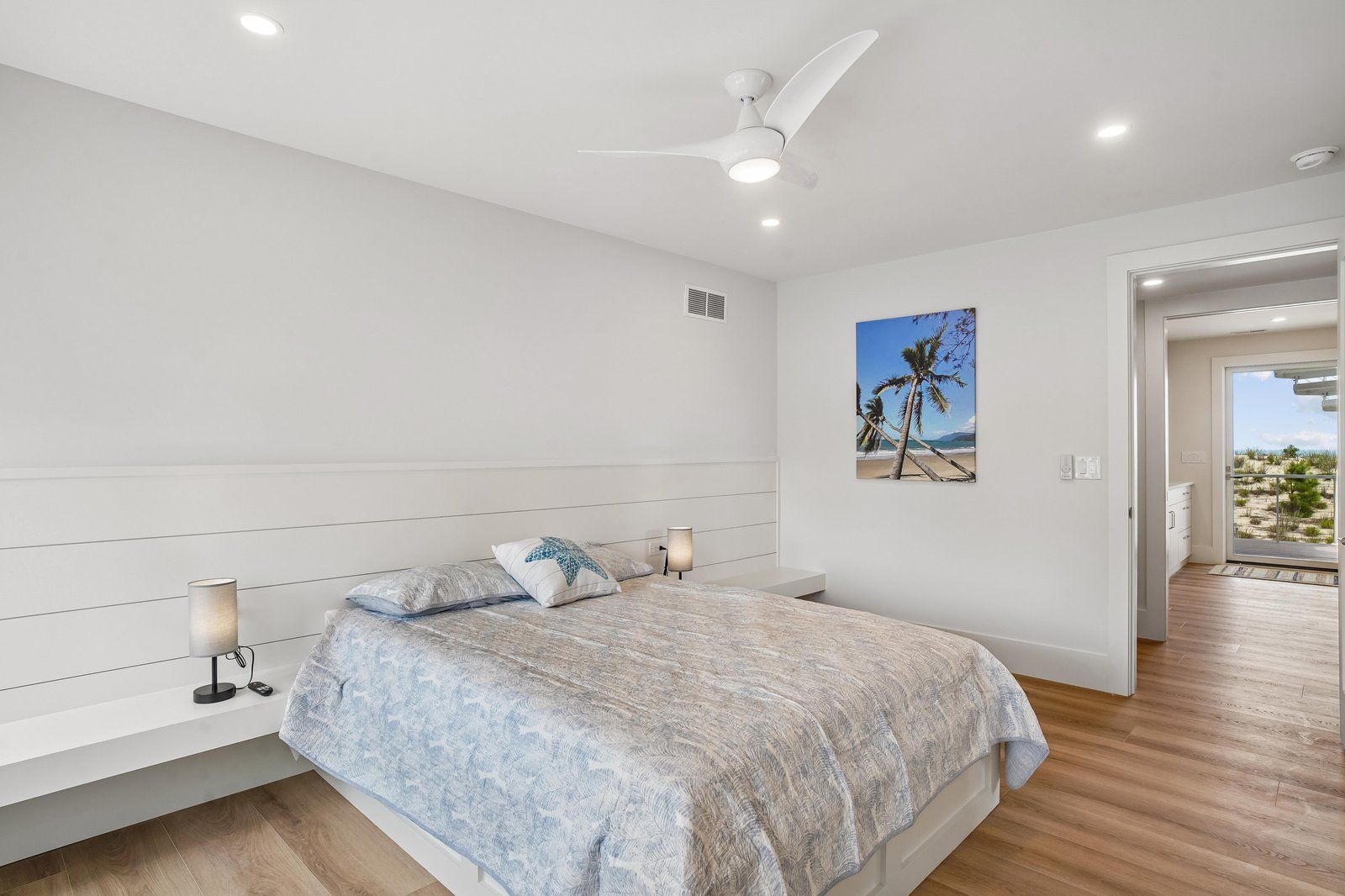 Un dormitorio con cama, lámparas de noche, ventilador de techo, conducto de aire en la pared y puerta que da a un balcón con vista a la playa.