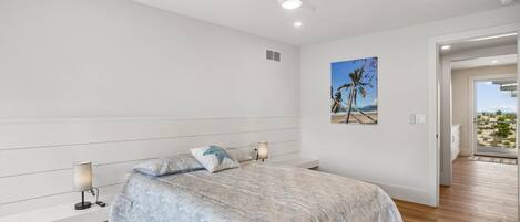 Un dormitorio con cama, lámparas de noche, ventilador de techo, conducto de aire en la pared y puerta que da a un balcón con vista a la playa.