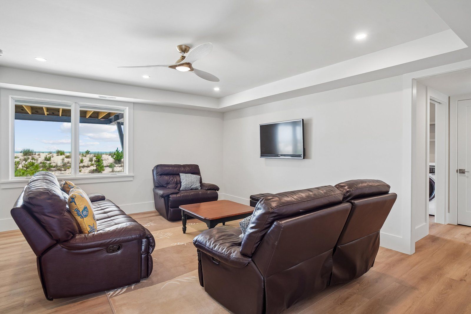 Un salon avec des canapés en cuir, un téléviseur à écran plat, un ventilateur de plafond et une vue sur la plage grâce à de grandes fenêtres.