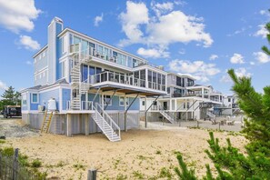 A multi-story beachfront property with a staircase leading to the upper levels, a sandy beach in the foreground, and a clear blue sky.