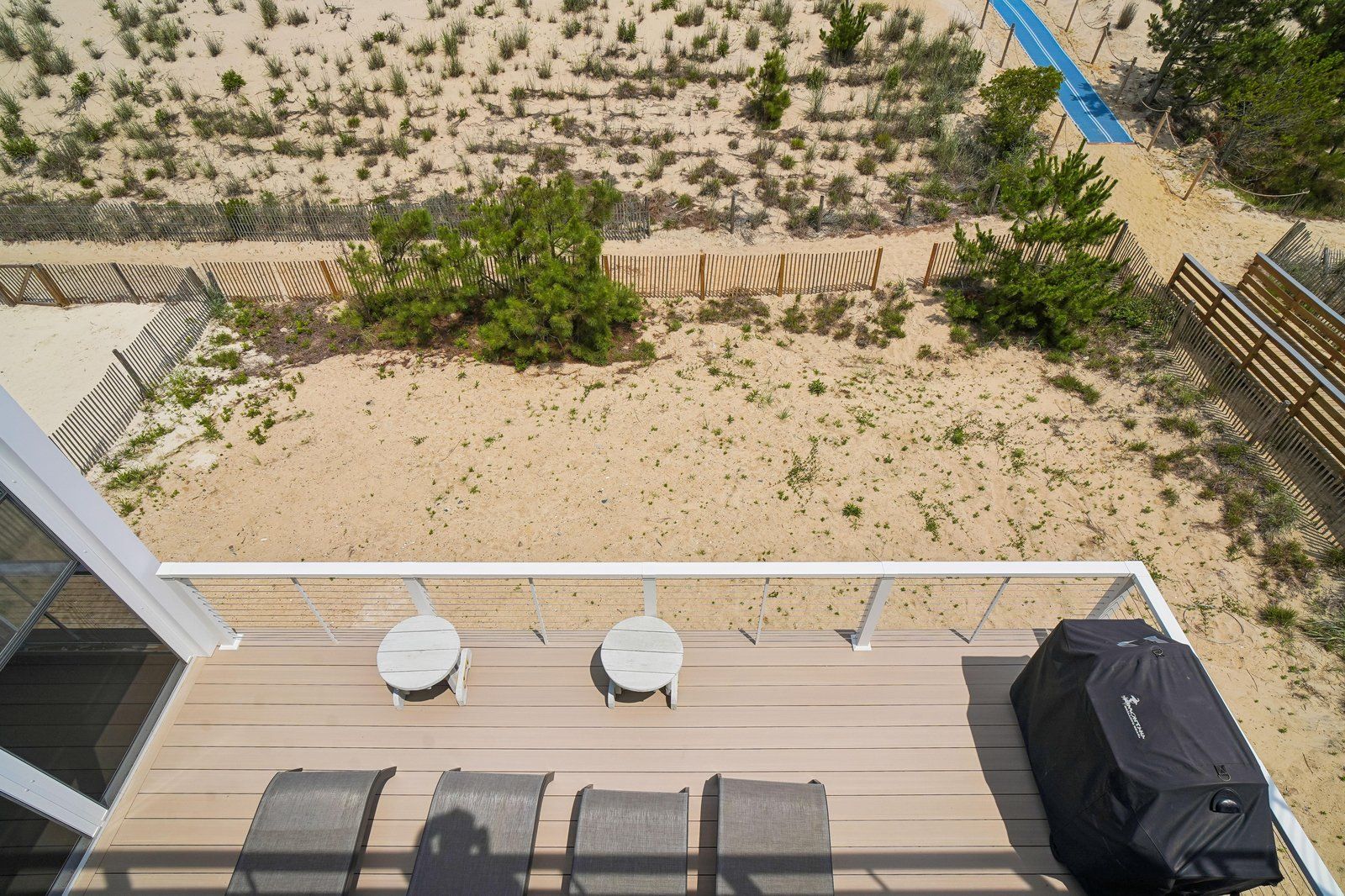 A deck with two tables and a grill, overlooking a sandy beach with sparse vegetation and a blue path.