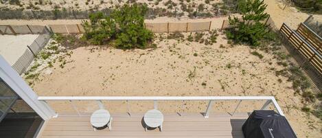 A deck with two tables and a grill, overlooking a sandy beach with sparse vegetation and a blue path.