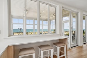 A room with a large window overlooking the beach, a white bench, and a telescope.