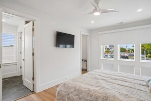 A bedroom with a bed, a ceiling fan, a TV, and a window with a view of a parking lot.