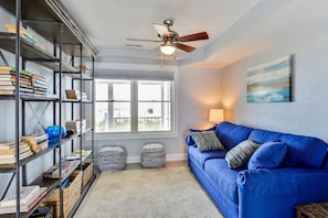 Ein Wohnzimmer mit einem blauen Sofa, einem Bücherregal, zwei Poufs, einem Deckenventilator und einem Fenster mit Blick.
