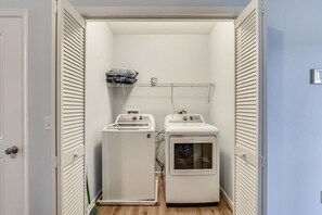 A laundry room with two washing machines and a drying machine, a shelf with folded clothes, and a door leading to another room.