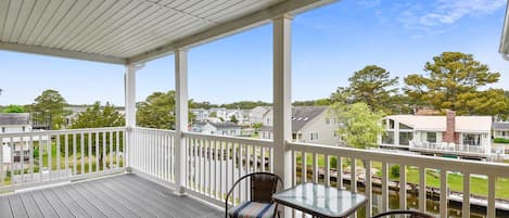 A covered outdoor deck with a glass table and chairs, overlooking a residential area with houses and a body of water.