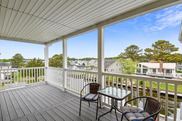 Um terraço coberto ao ar livre com mesa e cadeiras de vidro, com vista para uma área residencial com casas e um corpo d'água.