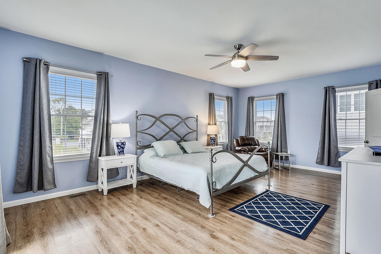 A bedroom with a bed, nightstands, a desk, a ceiling fan, and large windows with curtains.