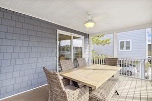 A covered patio with a wooden table and wicker chairs, a ceiling fan, and a view of a residential building.