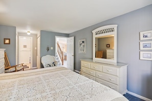 A bedroom with a large bed, a dresser, a chair, and a door leading to a staircase.