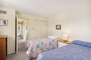 Habitación de hotel con dos camas, una mesita de noche de madera, una lámpara, un cuadro enmarcado en la pared y una puerta que da a otra habitación.