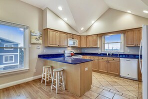 Une cuisine moderne avec des meubles en bois, un plan de travail bleu, un îlot central avec des tabourets et une vue sur une maison bleue par la fenêtre.