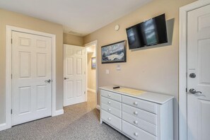 Un couloir avec deux portes blanches, une commode blanche à tiroirs, un téléviseur à écran plat fixé au mur et un tableau représentant un paysage, encadré, qui orne le mur.