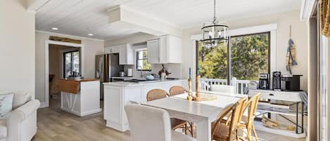 Cocina moderna con armarios blancos, una isla central y un comedor con mesa blanca y sillas de mimbre.