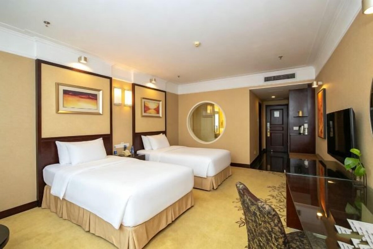 A hotel room with two beds, a flat-screen TV, a round mirror, and a carpeted floor.