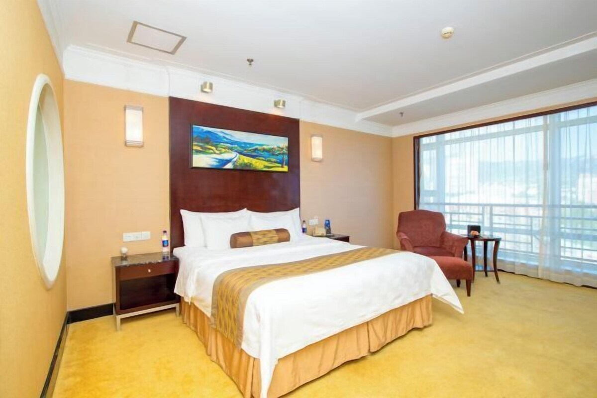 A hotel room with a large bed, a nightstand, a chair, and a large window.