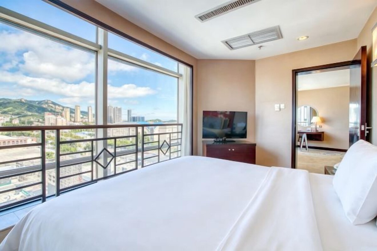 A hotel room with a large bed, a television, a desk, and a balcony with a city view.