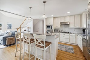 A modern kitchen with white cabinetry, a central island with bar stools, a stainless steel refrigerator, and a microwave. - Coastal Bliss Nestled in Bethany Beach: Oceanside, Elevator, Sleeps 16 (South Bethany)