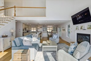 A spacious living room with a high ceiling, a staircase, a kitchen, and a fireplace. - Coastal Bliss Nestled in Bethany Beach: Oceanside, Elevator, Sleeps 16 (South Bethany)