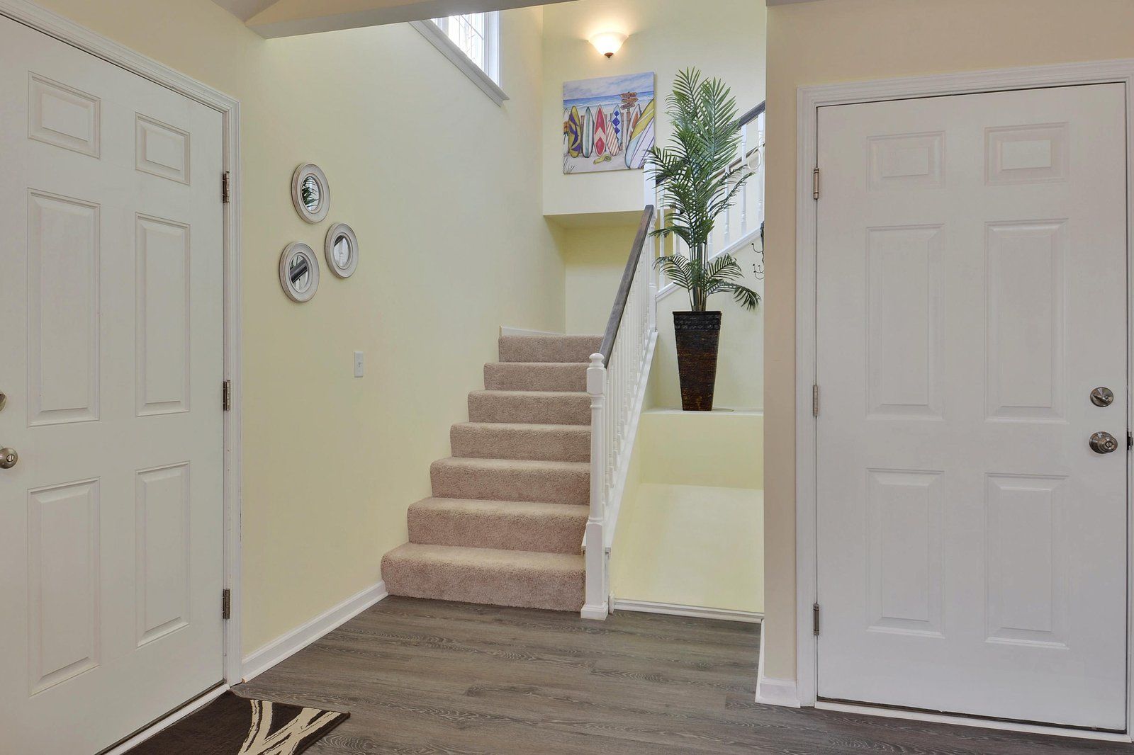 A staircase with carpeted steps leading to a landing, flanked by white doors, a wall-mounted light fixture, and a potted plant.