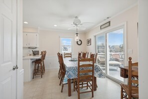 Uma sala de jantar com uma mesa posta para quatro pessoas, cadeiras de madeira, um ventilador de teto e vista através de grandes janelas.