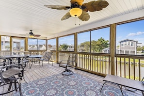 A covered outdoor patio with a table and chairs, a ceiling fan, and large windows overlooking a residential area. - Bayside South Bethany Home (South Bethany)
