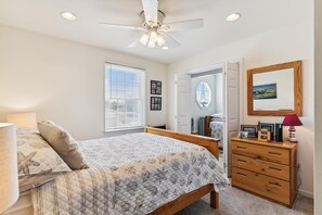 A bedroom with a bed, a dresser, a ceiling fan, a window with blinds, and a door leading to another room. - Bayside South Bethany Home (South Bethany)
