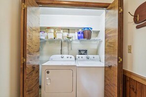 A laundry room with two white Whirlpool washers and dryers, shelves above holding various items including a bottle of Glidden, a blue container, and a brown bucket.