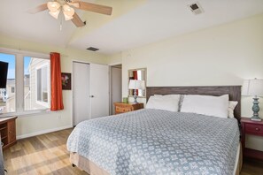 A bedroom with a bed, a nightstand, a dresser, a window with curtains, a ceiling fan, and a door. - The Buffer Zone: Oceanfront Bliss in Bethany Beach (South Bethany)