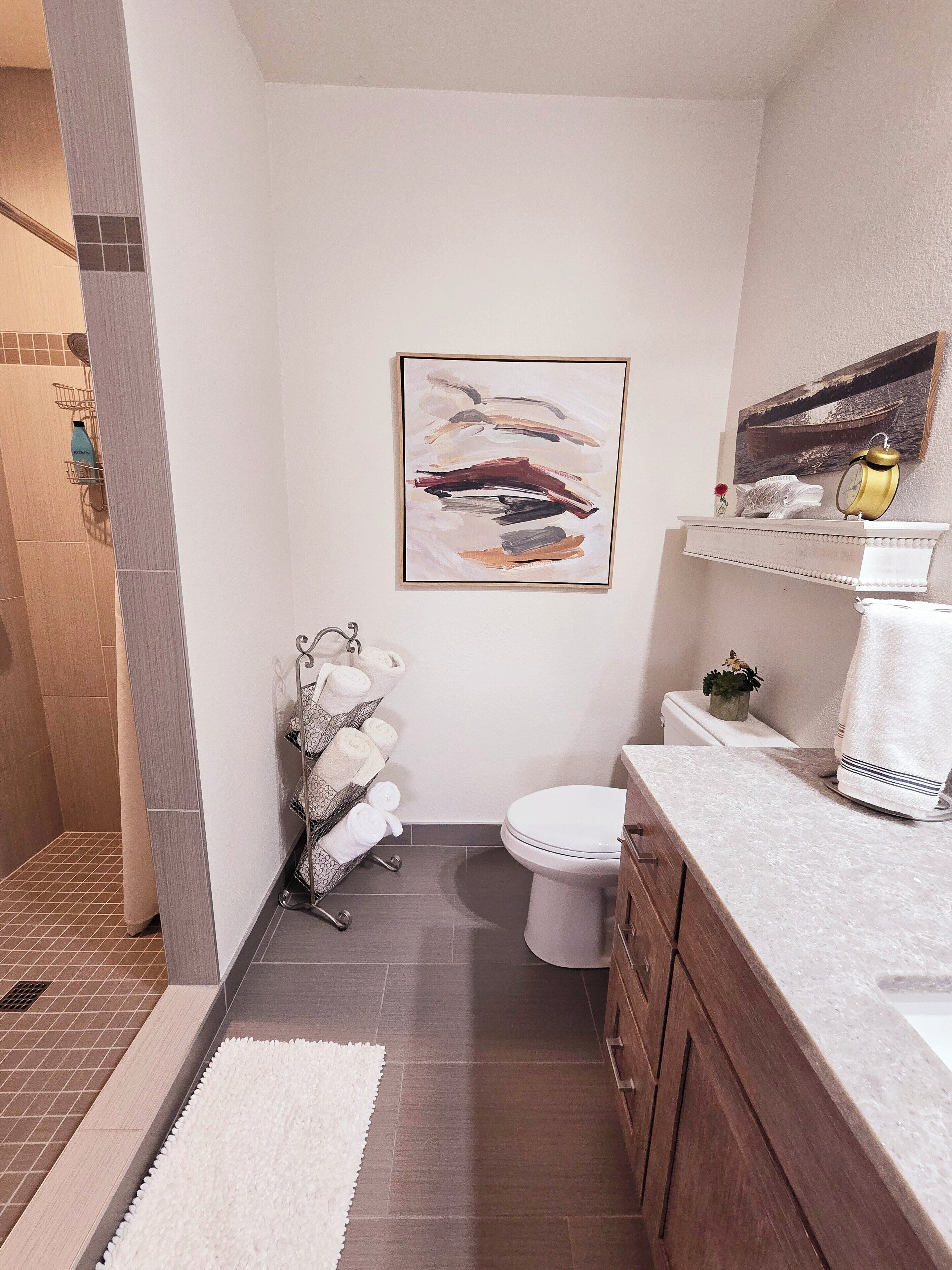 A modern bathroom with a shower, toilet, and sink. There is a towel rack with towels, a framed abstract painting, and a small potted plant on the vanity.