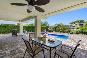 En udendørs terrasse med et glasbord, stole, en ventilator i loftet, grill og en swimmingpool i baggrunden.