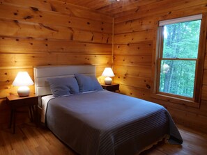 Un dormitorio de cabaña de madera con una cama, dos lámparas de noche y un ventanal con vista al bosque.