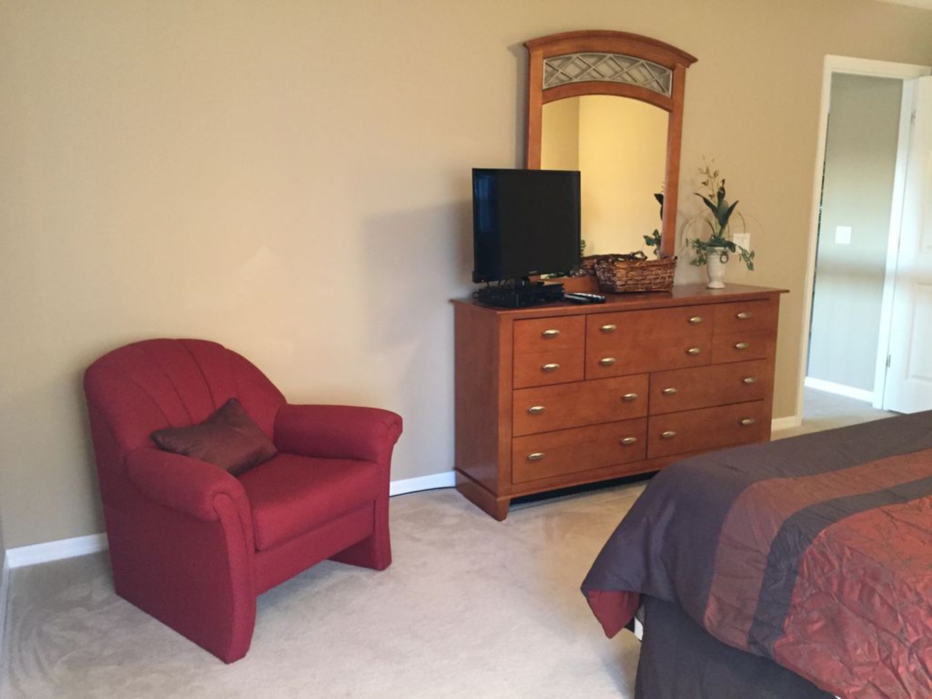 Une chambre à coucher avec un fauteuil rouge, une commode en bois dotée d’un miroir et un lit recouvert d’une couverture de couleur sombre.