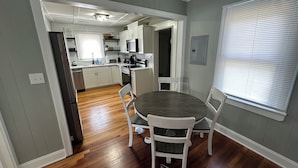 A dining area with a round table and four chairs, a kitchen in the background, and a window with blinds. - City Cottage Winston-Salem
 (Winston-Salem)