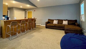 A living room with a ceiling fan, a brown sofa, a blue bean bag, and a kitchen area with bar stools.