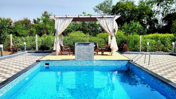 A swimming pool with a tiled edge and a logo in the center. There are two wooden chairs under a white canopy. - Teakwood Farms (Wardha)