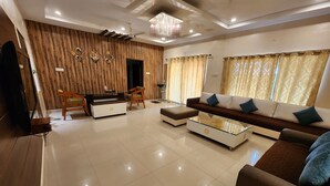 A modern living room with a wooden accent wall, a sofa set, a glass coffee table, and a dining area with a wooden table and chairs. - Teakwood Farms (Wardha)