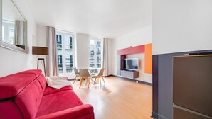 Una sala moderna con un sofá rojo, una mesa de comedor con sillas, un televisor y un amplio ventanal con vista a edificios.
