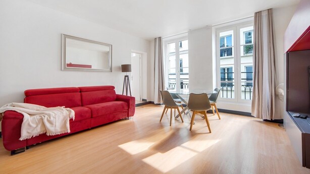 Una sala de estar moderna con un sofá rojo, una mesa de comedor de cristal con sillas, una lámpara de pie y ventanas grandes que ofrecen vistas al exterior.