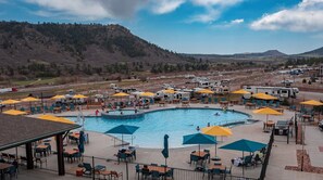 Zona de piscina con varias sombrillas y diferentes espacios de descanso, rodeada de vehículos recreativos y montañas al fondo.