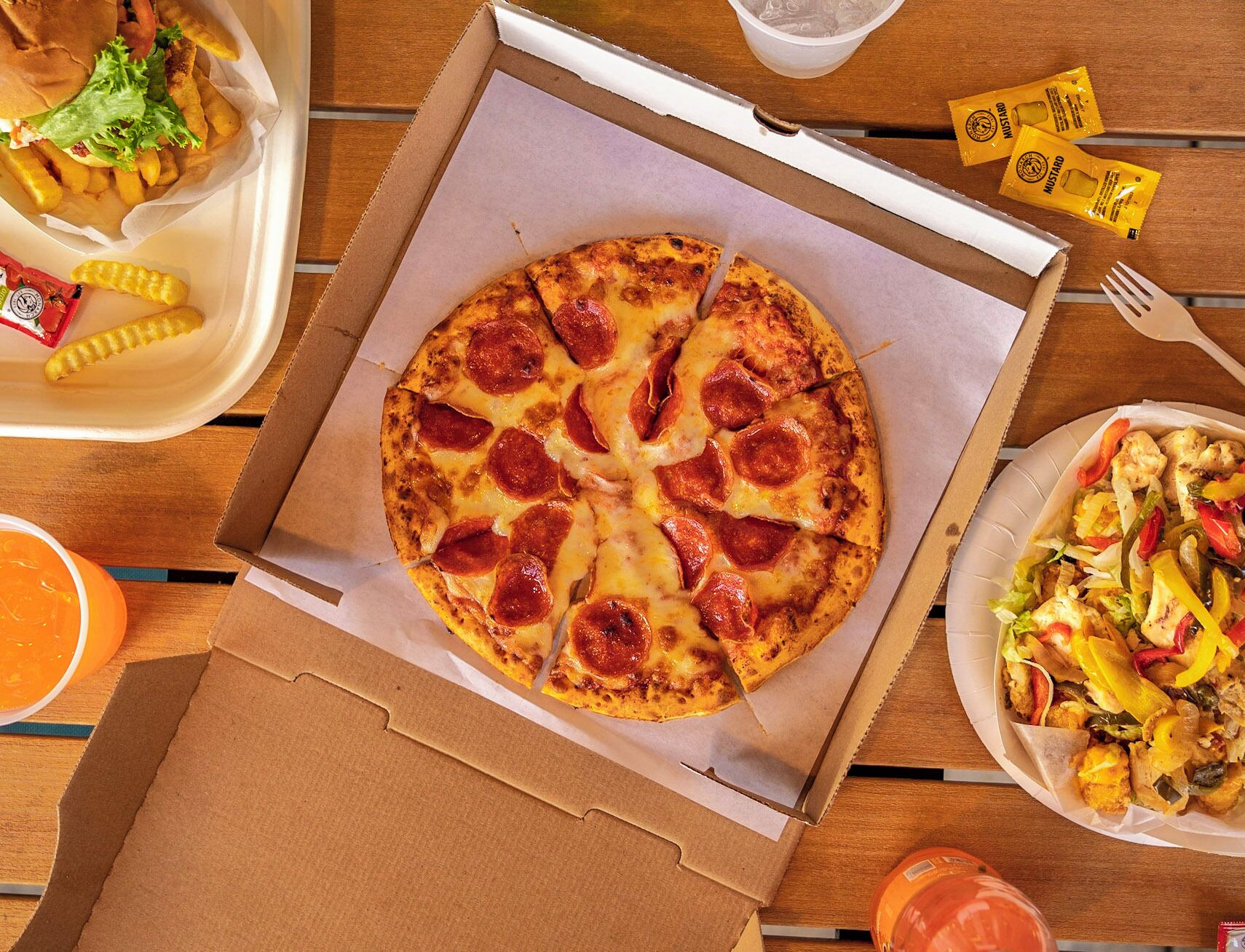 Una pizza en una caja de cartón con lonzas de pepperoni, una porción de papas fritas, una ensalada y salsas en una mesa de madera.
