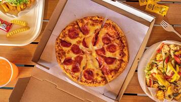Una pizza en una caja de cartón con lonzas de pepperoni, una porción de papas fritas, una ensalada y salsas en una mesa de madera.