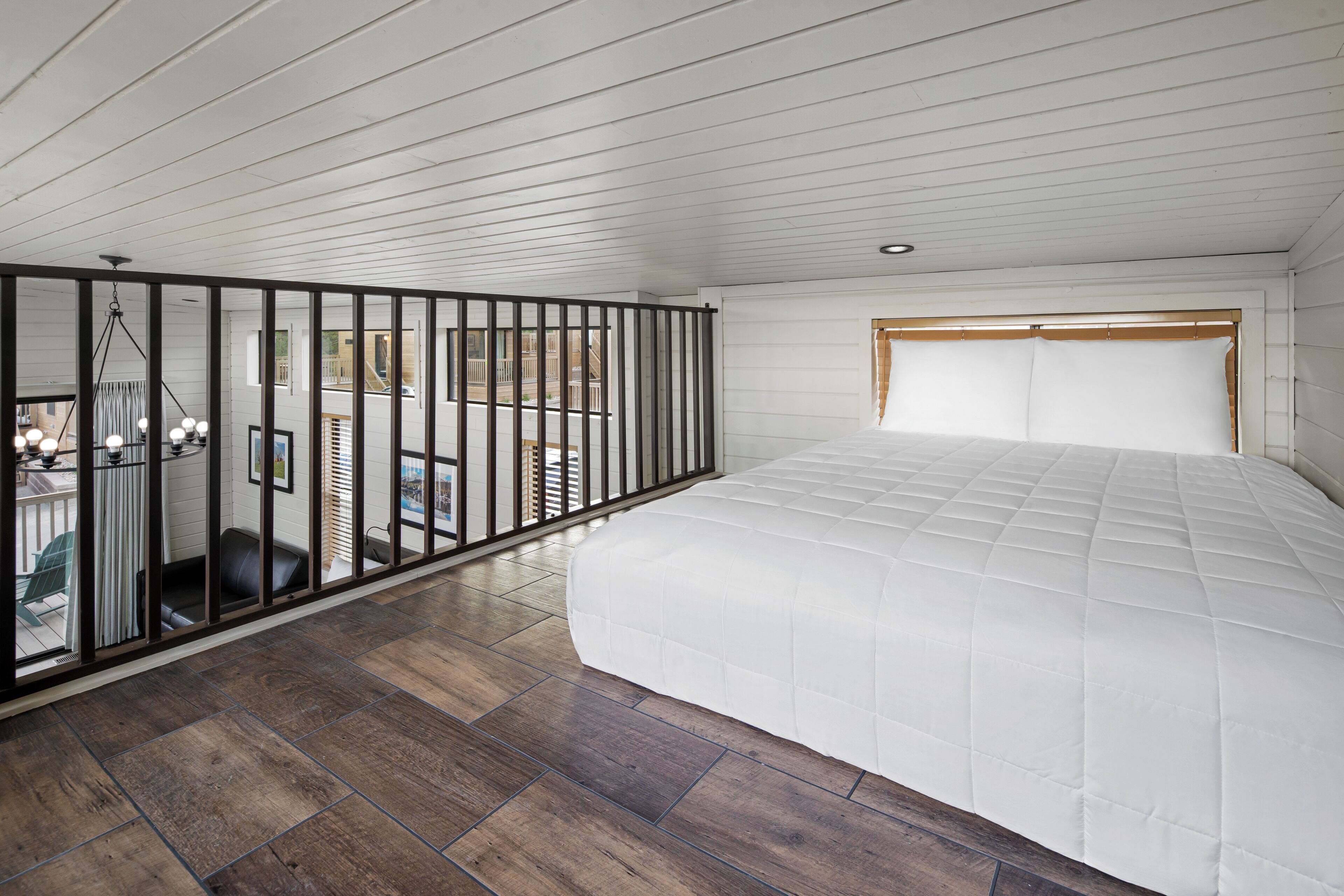 A bedroom with a large bed, wooden flooring, a balcony with a railing, and a view of the outside.