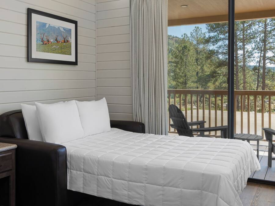 A bedroom with a bed, a nightstand, a chair, and a balcony with a view of trees and mountains.
