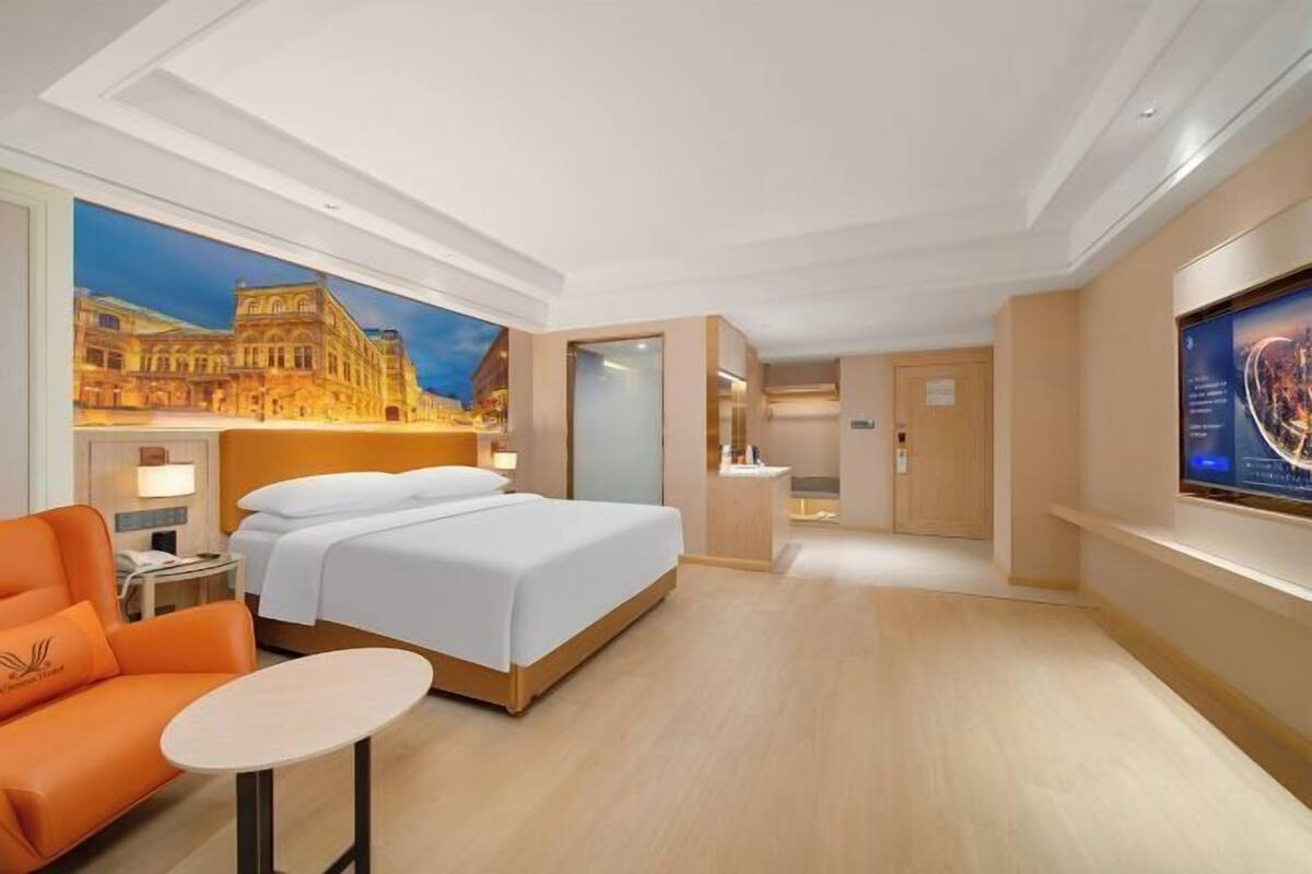 A hotel room with a large bed, a small table, an orange chair, a bathroom, and a TV screen.