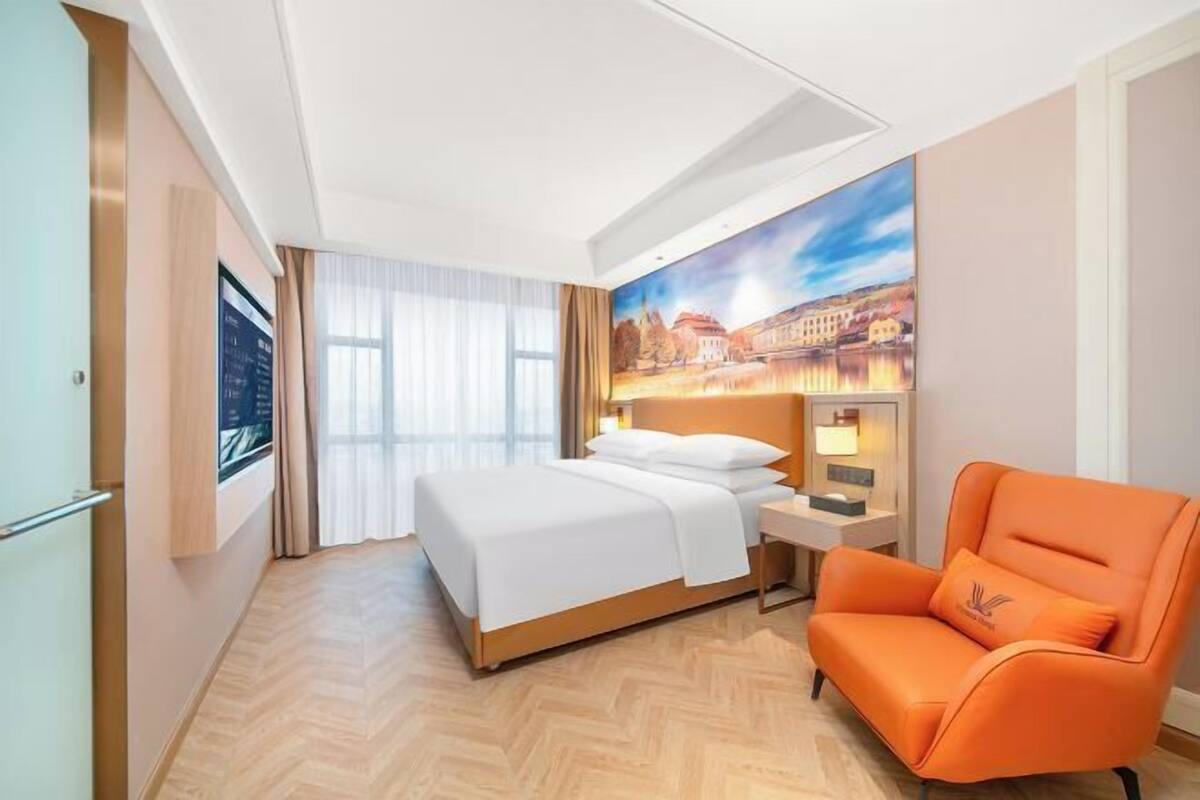 A hotel room with a bed, a nightstand, a chair, a painting on the wall, and a window with curtains.
