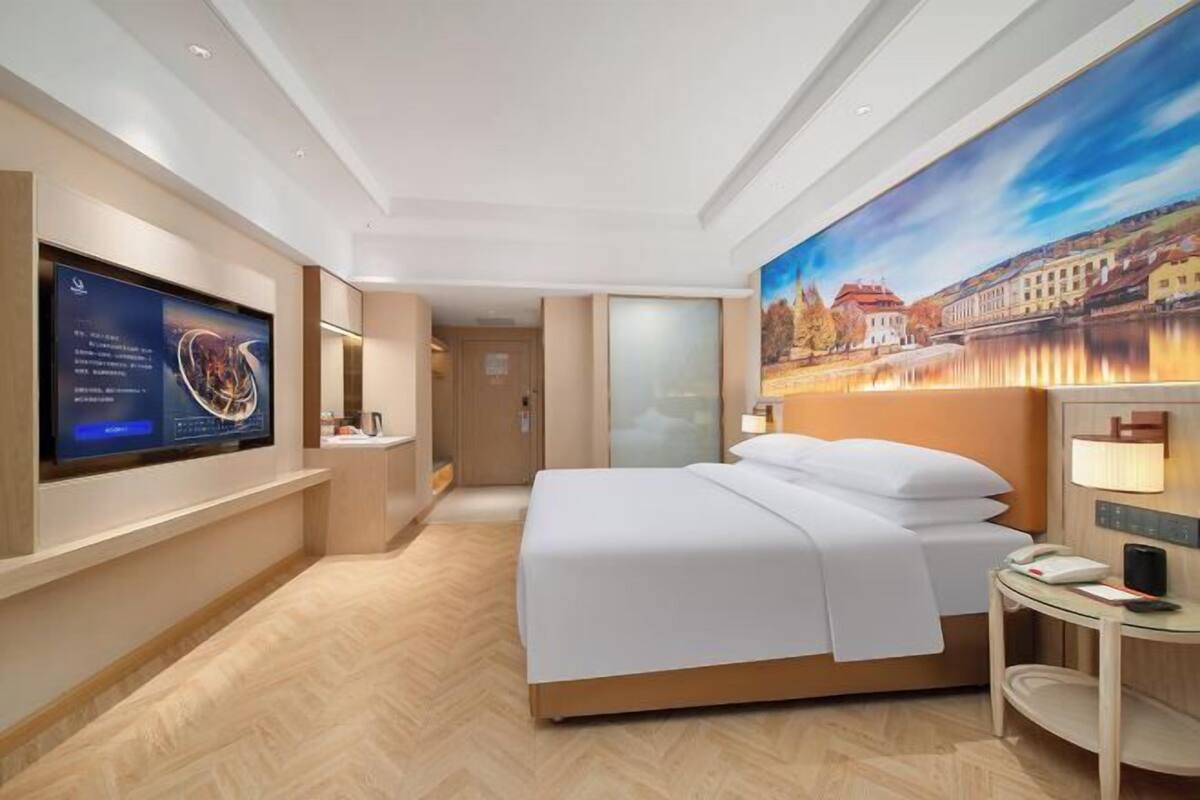 A modern hotel room with a large bed, a flat-screen TV mounted on the wall, a painting of a scenic landscape, and a small table with a lamp.